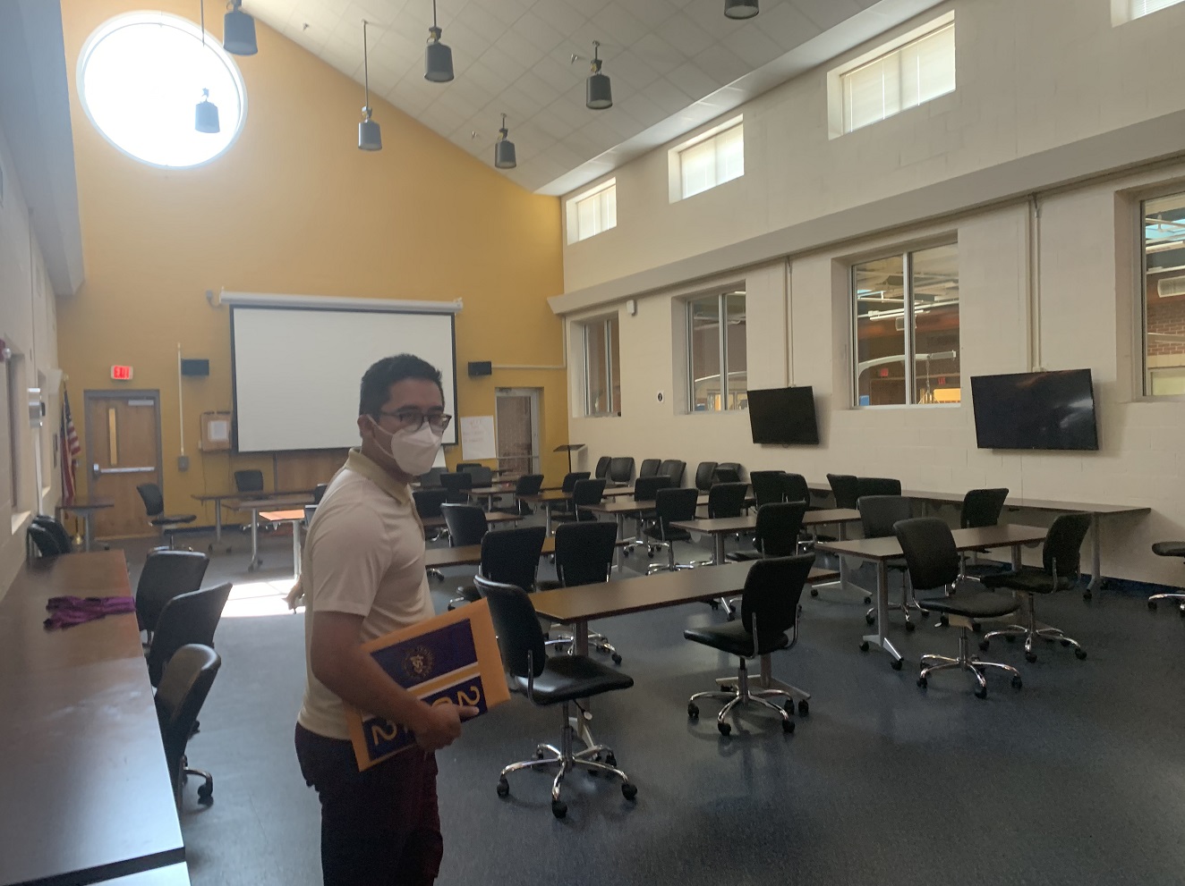 A man with short dark hair and a white t-shirt and face mask stands in front of a room filled with long tables and rolling chairs. In the front of the room is a projector screen, and there are two large monitors on the side wall. There are windows into another indoor space, plus higher windows to let in natural light, including a large round window at the peak of the sloped ceiling.